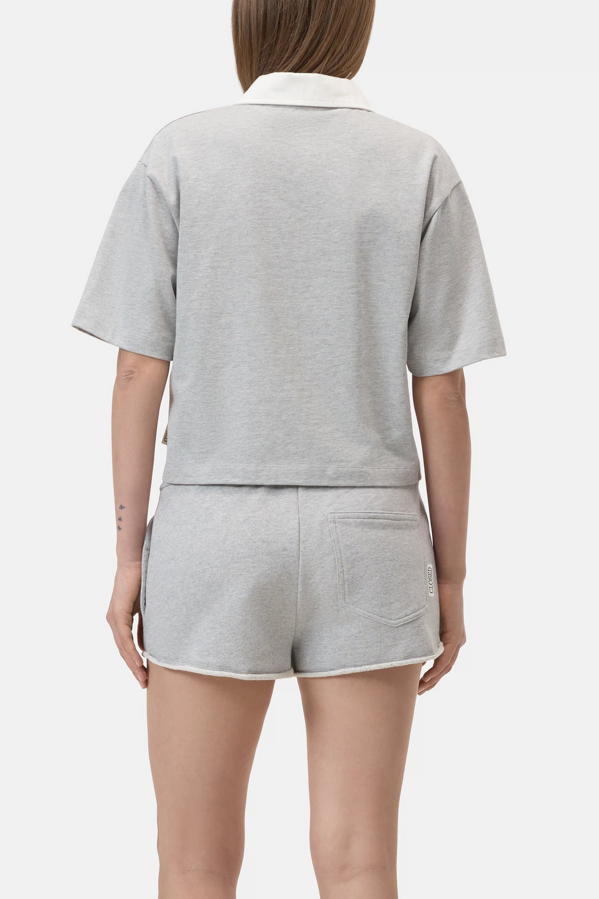 A person is shown from behind wearing the CLOSED Cropped Rugby Shirt in light gray with a white collar, paired with matching shorts against a plain white background.