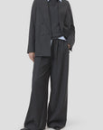 A person wears CLOSED’s Fitted Doublebreasted Blazer in dark gray with wide-leg trousers over a light blue collared shirt, standing against a plain light background. Their face is partially out of frame.