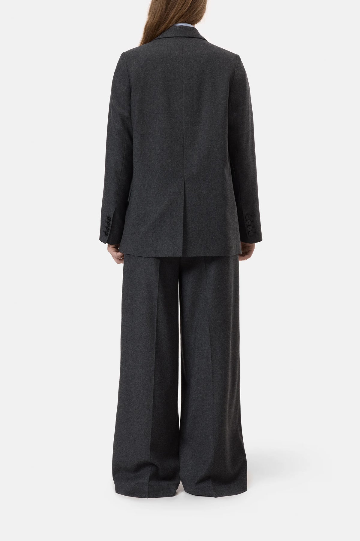 A person with long brown hair stands facing away against a plain white background, wearing CLOSED’s Fitted Doublebreasted Blazer in dark gray paired with wide-leg trousers.