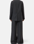 A person with long brown hair stands facing away against a plain white background, wearing CLOSED’s Fitted Doublebreasted Blazer in dark gray paired with wide-leg trousers.
