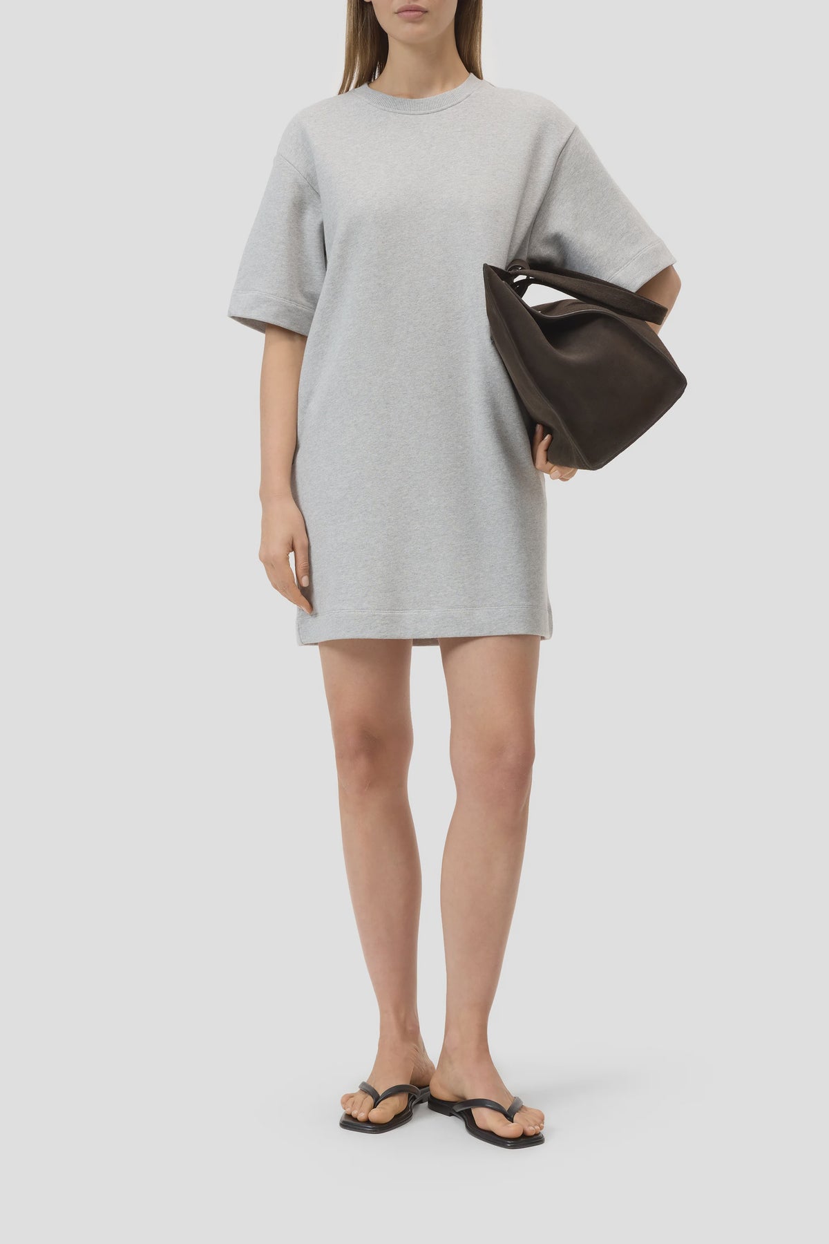 A woman stands against a plain light background in a loose, short-sleeved, light gray CLOSED Felpa Dress (100% cotton), black flip-flop sandals, and holds a large dark brown handbag under her arm.