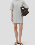 A woman stands against a plain light background in a loose, short-sleeved, light gray CLOSED Felpa Dress (100% cotton), black flip-flop sandals, and holds a large dark brown handbag under her arm.