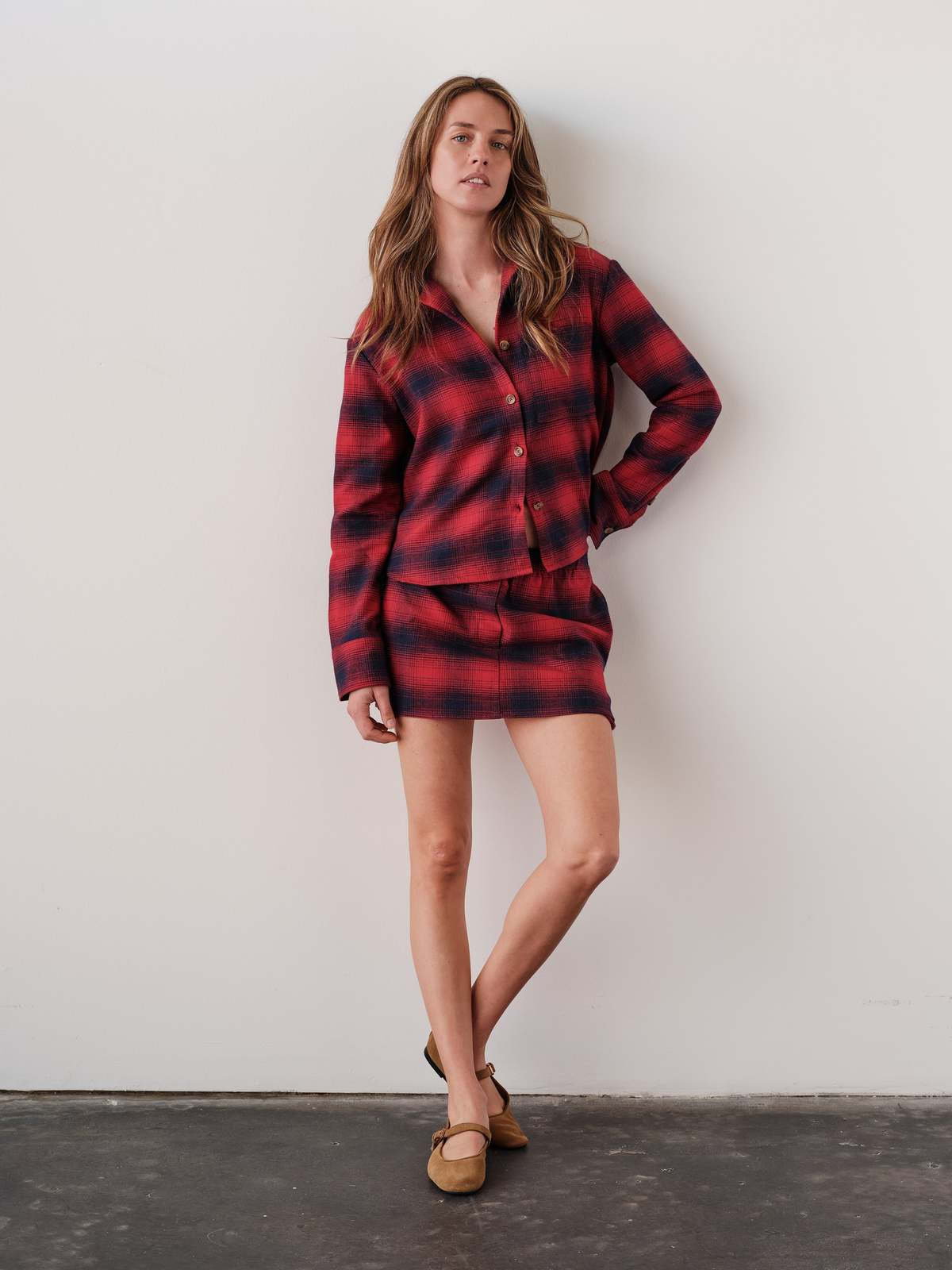 The MINI Skirt in a red plaid pattern, styled with tan loafers, modeled by Cissa against a white wall.