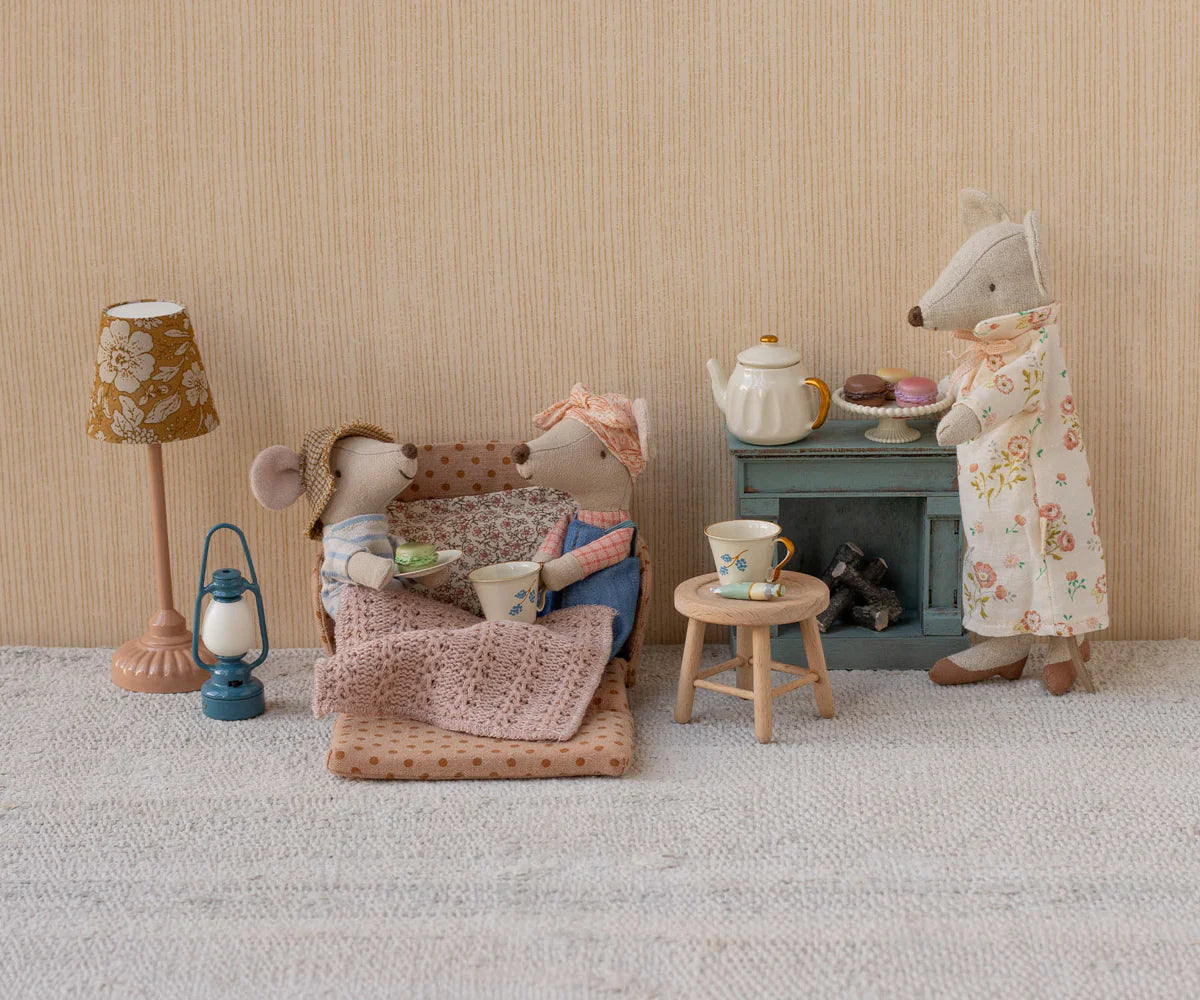 Two Maileg mice lounge on the Maileg Sofa Bed with tea cups and a pink blanket, while a third mouse serves tea and pastries by a blue cabinet. The charming scene also includes polka-dot accents, a lamp, and a small lantern.