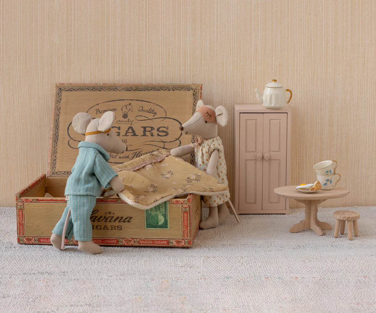 Maileg Mum & Dad Mice in a cigar box bed—one wears pajamas, the other a dress and bow. Nearby are a small cabinet, round table with teapot and cake, and a tiny wooden stool. All by Maileg.