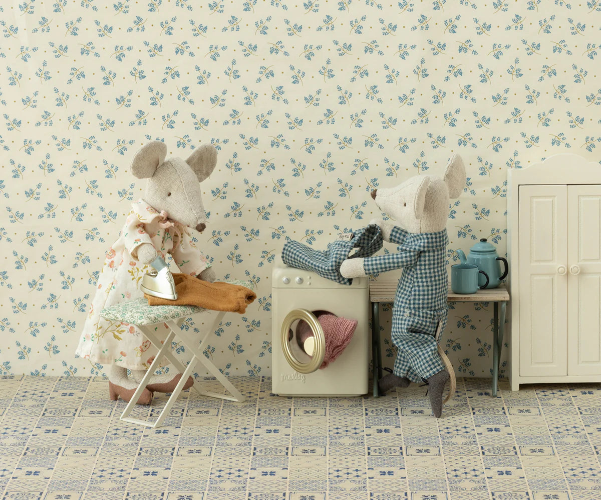 Two Maileg mouse figures—a dress-wearing one ironing and a pajama-clad one loading laundry—enjoy the Maileg Washing Machine in a cozy miniature room, complete with wardrobe, table, teapot, cups, and a floral patterned floor.