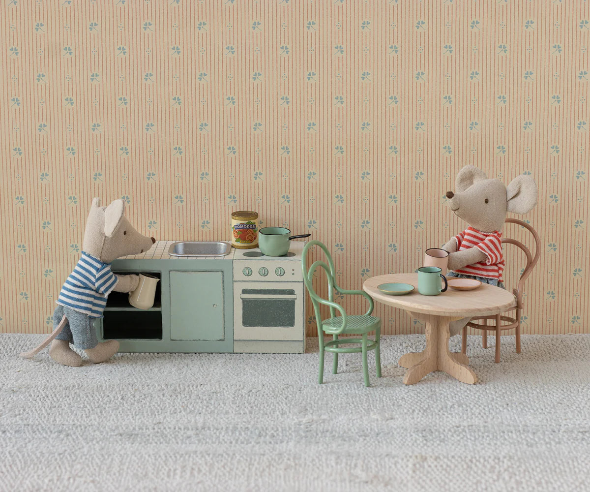 Two Maileg toy mice in striped shirts create a cosy dinner scene: one cooks at the stove, while the other sits at the Maileg Oval dining table. The miniature setting features patterned wallpaper and a textured floor, all by Maileg.