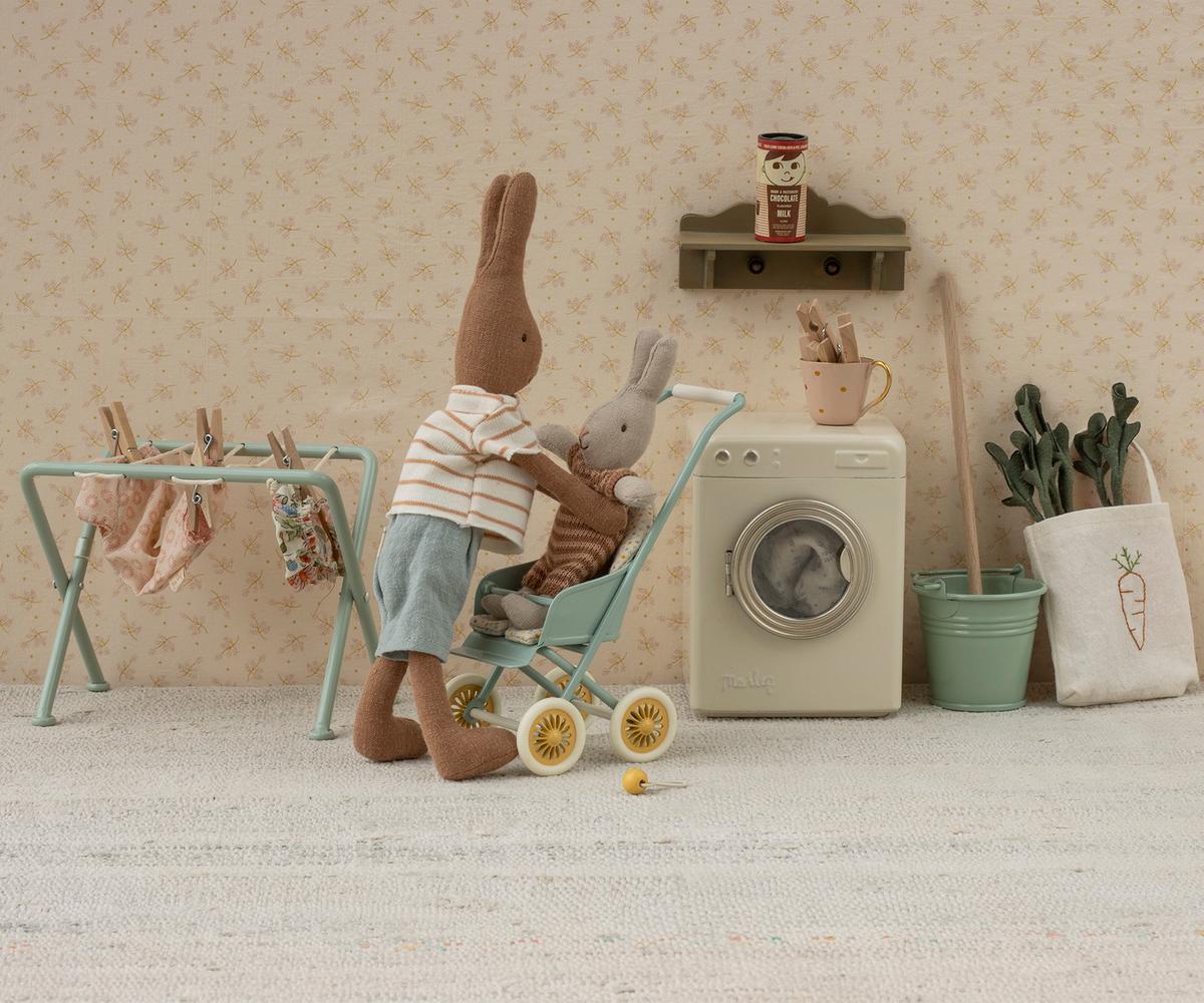 Two plush Maileg rabbits, one pushing the other in a toy stroller, stand in a miniature laundry room with a washing machine, drying rack, potted plants, and Maileg Carrots in Bag for a cute touch to imaginative play.