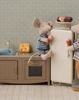 Two Maileg mice enjoy ice cream in the Kitchen set. One mouse sits on the counter, another stands by the open fridge. A plush fish, cheese wedge, and patterned wallpaper add charm to this delightful Maileg family scene.
