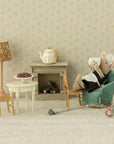 Maileg’s Big Sister Maid Mouse relaxes in an armchair, sipping tea in a miniature Maileg living room with a table, chairs, lamp, fireplace, teapot, pie, and a vacuum cleaner on the floor.