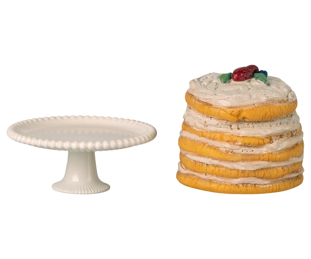 The Maileg Party Cake by Maileg is displayed on a white decorative cake stand beside pancakes with white icing and colorful fruit, all set against a plain background.