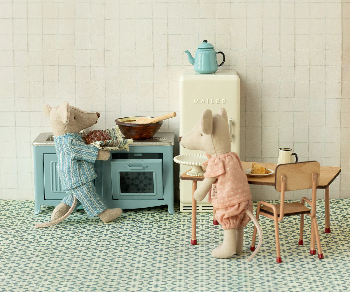 Two Maileg mice in pajamas enjoy their miniature Kitchen set, which features a blue stove, table with chairs and plate, small fridge, and blue teapot—all arranged on a tiled floor for delightful pretend play.