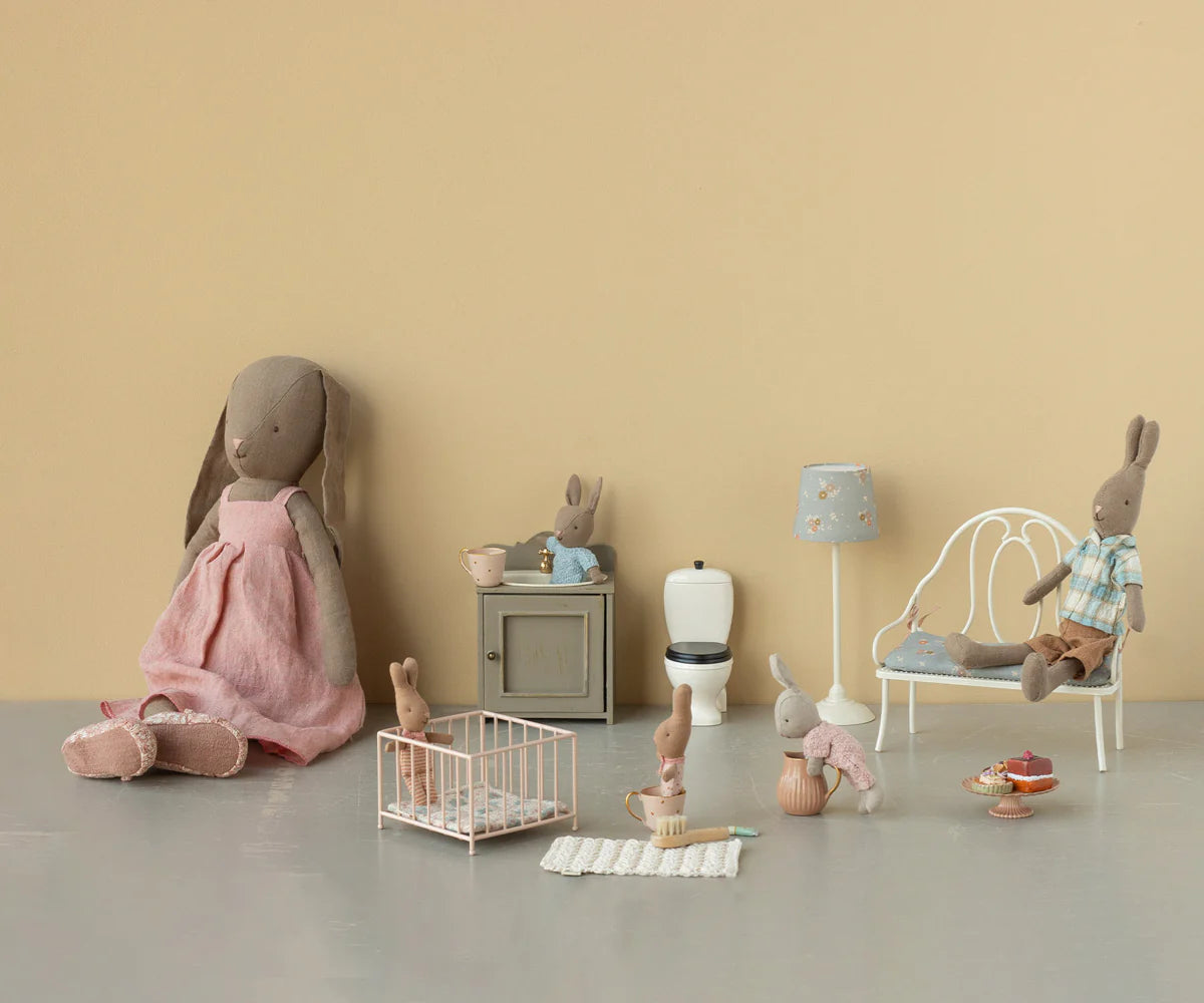 Maileg plush bunnies in different outfits are displayed in a miniature room with Maileg doll furniture, including the Maileg Playpen, crib, bed, cabinet, toilet, lamp, and tiny food items against a beige background.