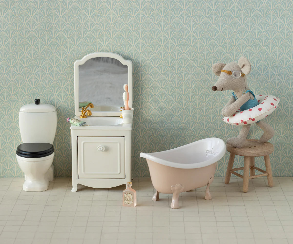 The Maileg Little Sister Beach Mouse by Maileg sits on a stool in a miniature bathroom with a toilet, sink, bathtub, and patterned wallpaper. She wears glasses, a swimsuit, and a swim ring for adorable beach-themed play.