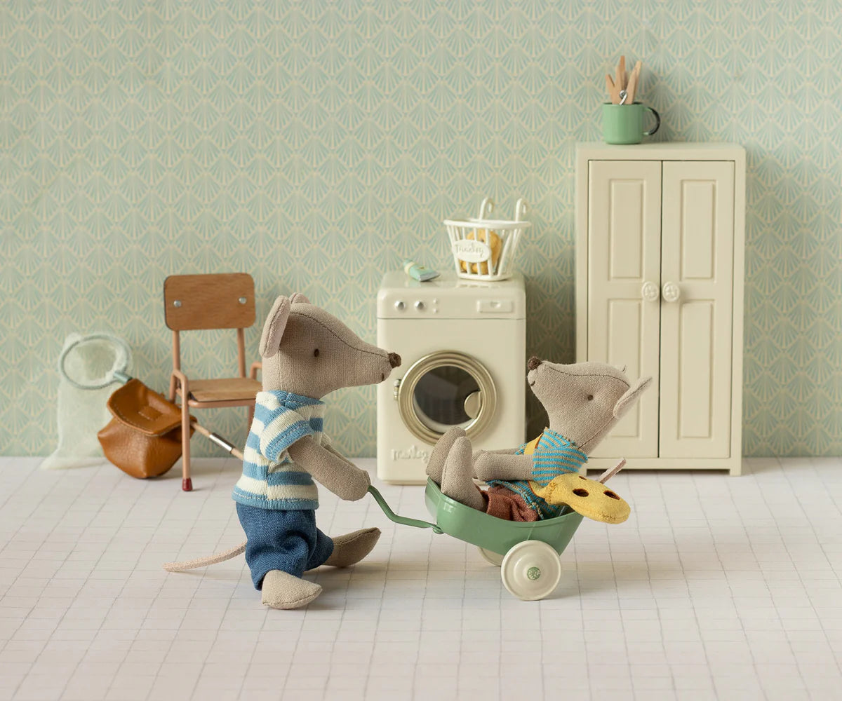 Little Brother Mouse with bag by Maileg pushes a wheelbarrow in a miniature laundry room, playful and charming scene.
