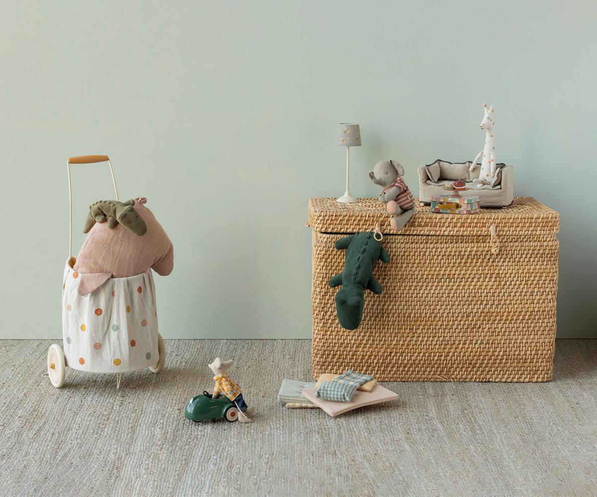 A wicker toy chest with plush animals, a toy lamp, and a miniature tea set stands near the Maileg Trolley by Maileg. Soft linen cushions and stuffed animals are scattered on the floor, creating a cozy and washable play space.