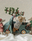 Three Maileg mice in festive winter outfits loading a Christmas tree onto a small blue toy truck.