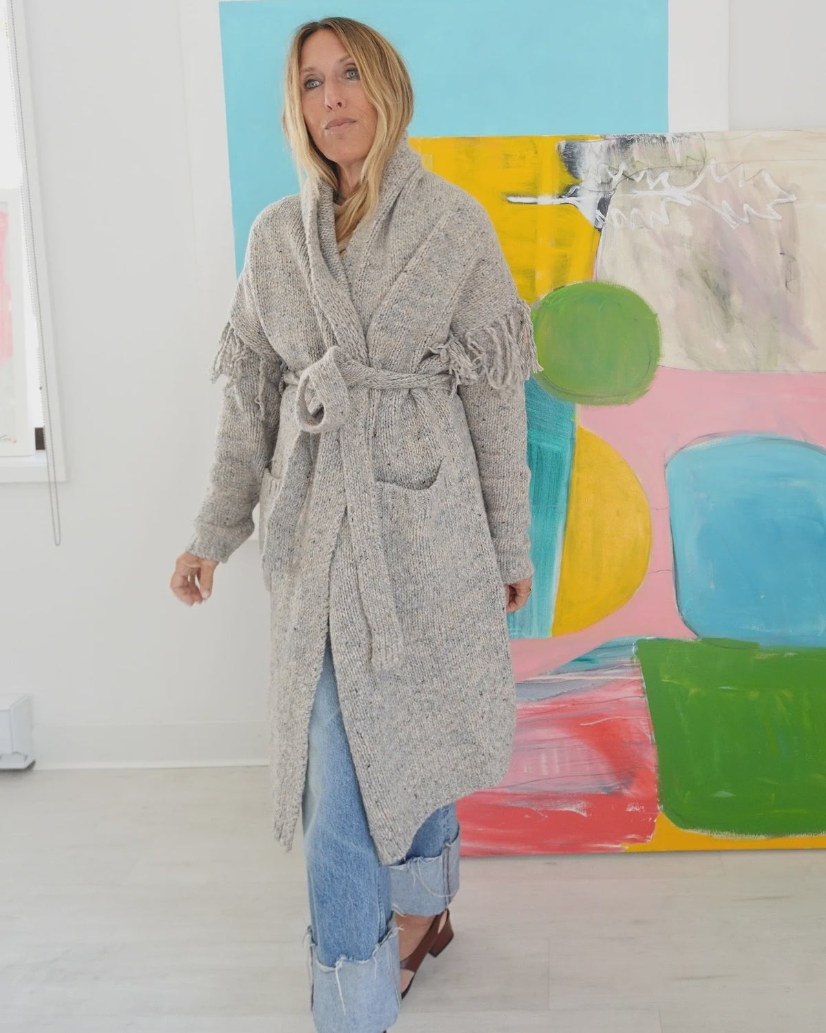 Cali Fringe Cardigan in grey merino wool with fringe shoulders and tie waist, styled over jeans.