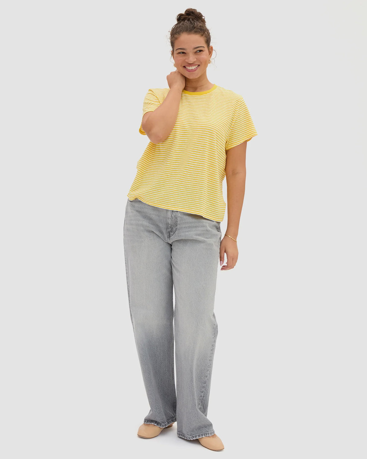 A person smiles, wearing the Clare Vivier Classic Tee Mini Stripe in yellow, paired with light blue wide-leg jeans and beige shoes, one hand touching their neck against a plain light background.
