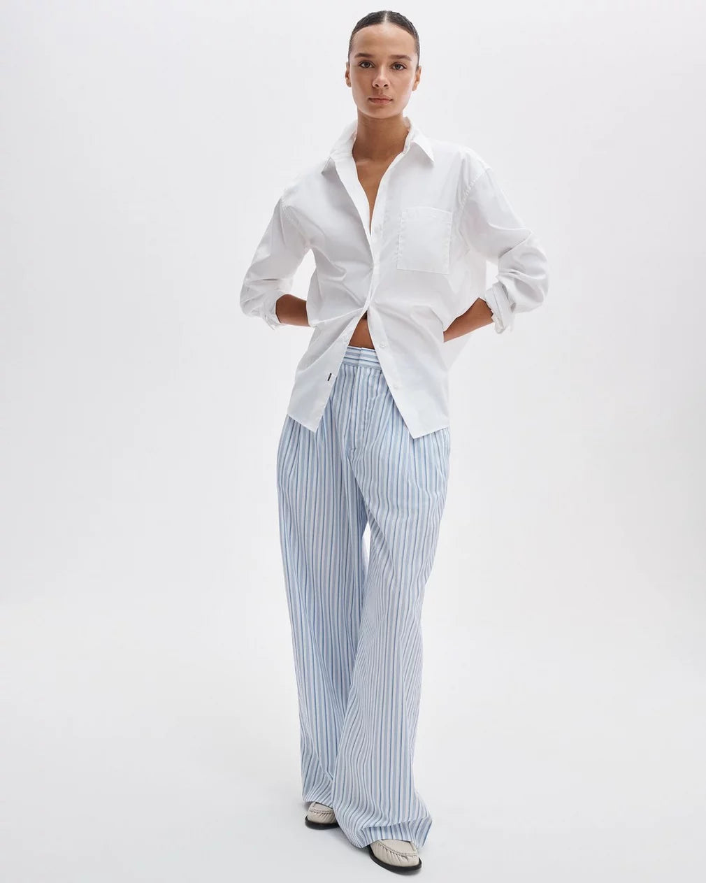 A person stands against a plain white background wearing the Rag & Bone connie shirt, a relaxed-fit cotton style with rolled sleeves, paired with light blue and white striped wide-leg pants and white shoes.