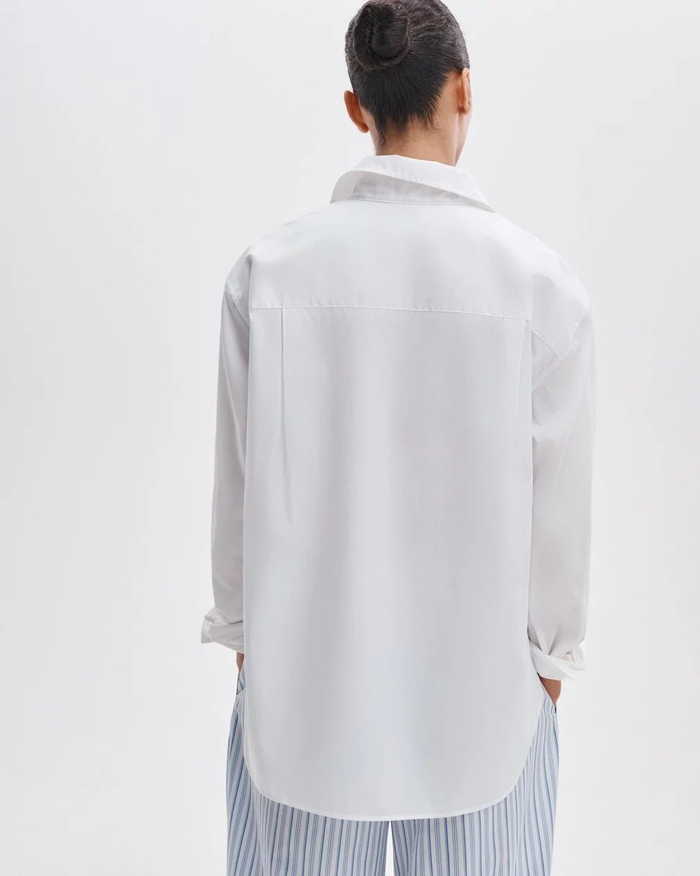 A person with dark hair in a bun, seen from behind, wears the Rag & Bone connie shirt and light blue and white striped pants while standing against a plain light background.