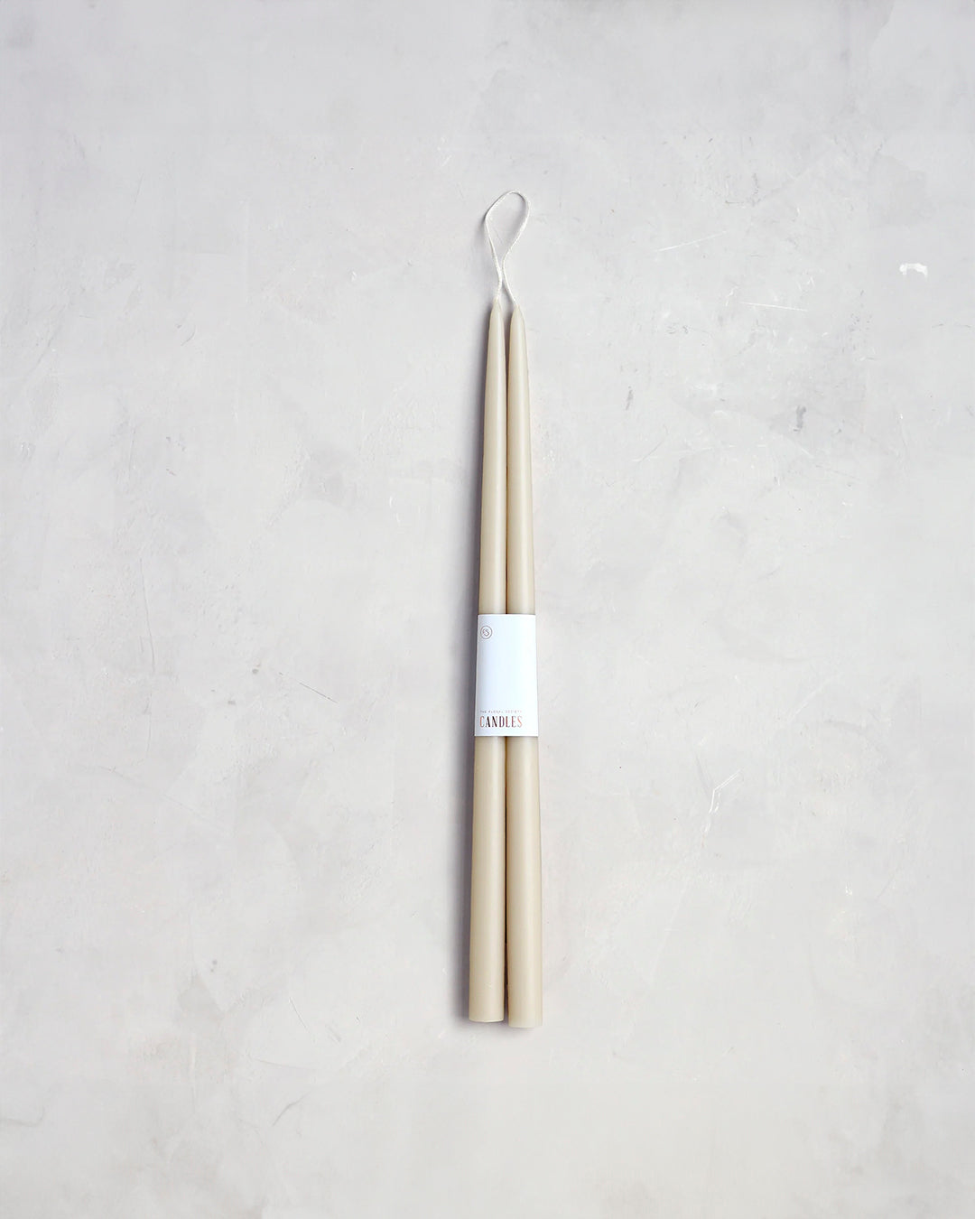 A pair of 18" Taper Candles from The Floral Society, bound together by a white label with minimalist text, exudes a classic design. Set against a light, textured background, these beige candles are attached at the wick ends by a white string loop, promising to bring a graceful ambiance to any setting.