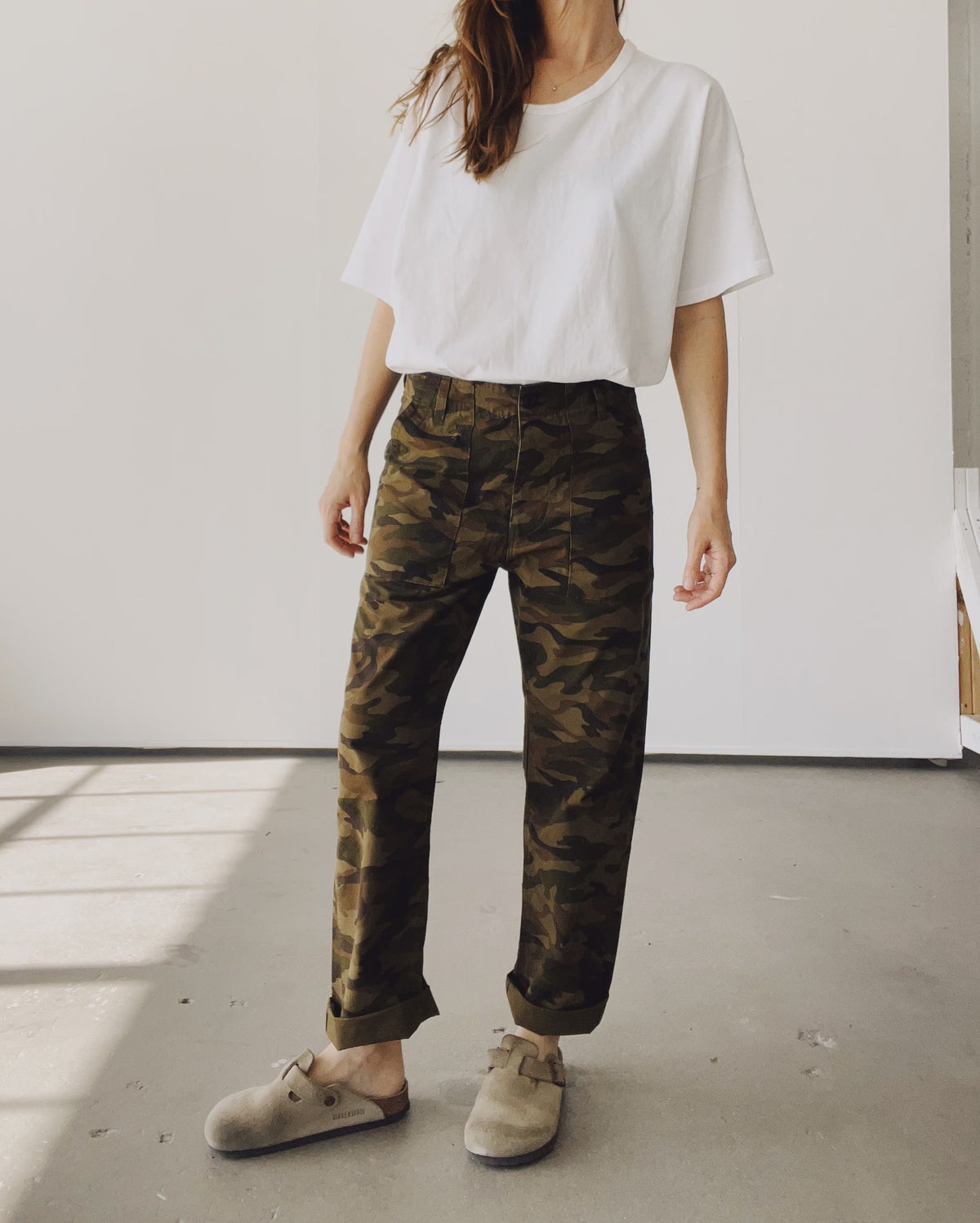 Someone stands indoors by a plain light wall, wearing a loose white tee, beige slip-on shoes, and Hey Gang’s The Officer Pant in 100% Cotton Ripstop with rolled cuffs. Their face is not visible.