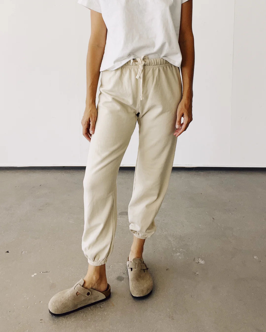 Standing on a concrete floor against a white wall, the person wears a white t-shirt, The Sweatpants by Hey Gang in beige French Terry with an elastic waistband, and light brown clogs.