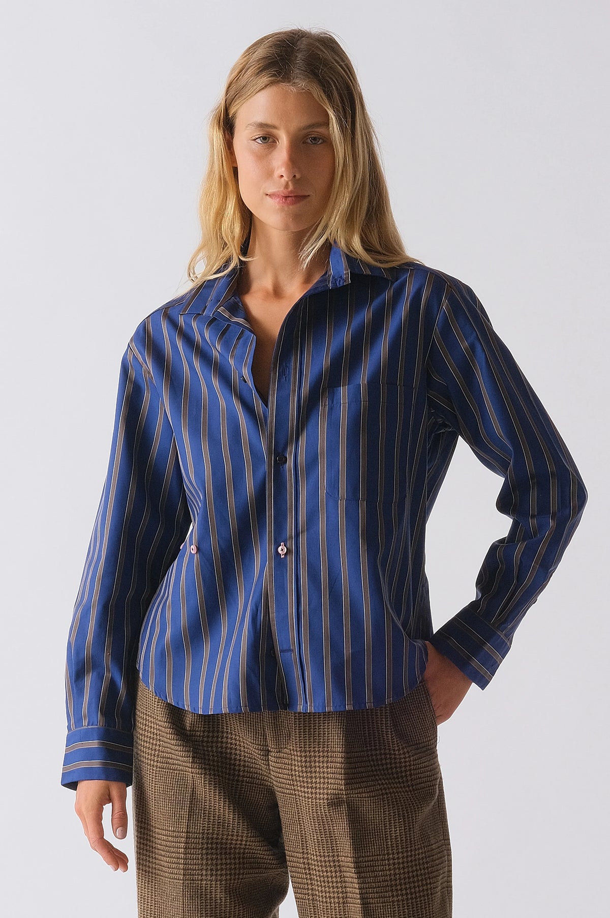 A person wears Cissa's The Daily Shirt—a blue PAPER COTTON button-up crop shirt with beige and white stripes—styled with brown plaid pants, standing against a plain light background, perfect for a daily look.