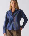 A person wears Cissa's The Daily Shirt—a blue PAPER COTTON button-up crop shirt with beige and white stripes—styled with brown plaid pants, standing against a plain light background, perfect for a daily look.