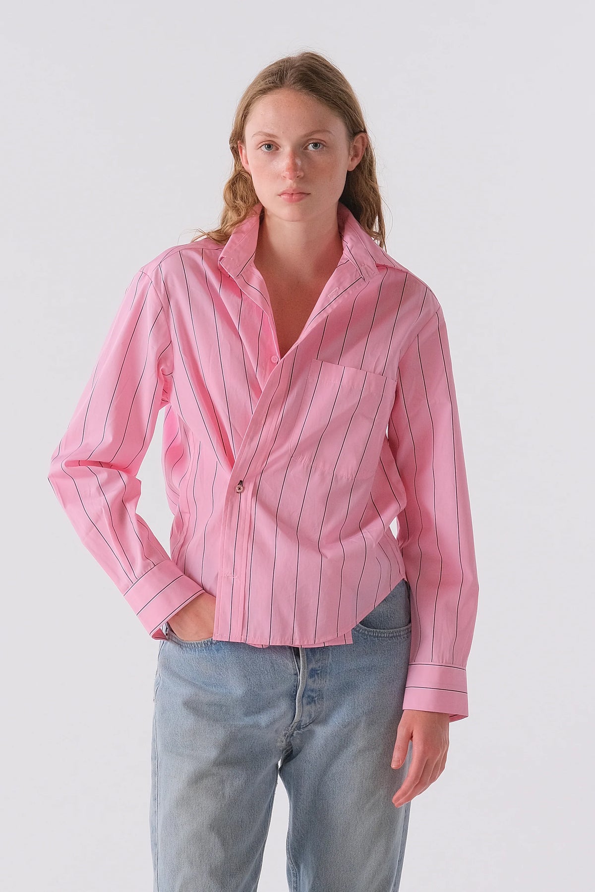 A person with long, light brown hair wears the Cissa Daily Shirt in pink with blue stripes and light blue jeans, posing with one hand in their pocket. Made from soft PAPER COTTON for all-day comfort, set against a plain light background.