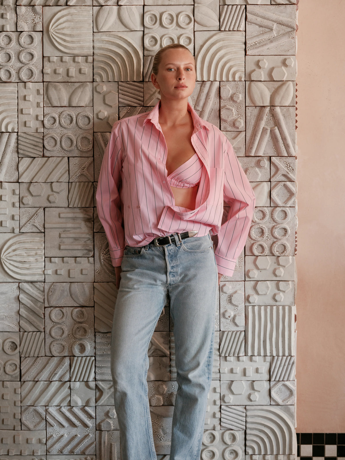 A person stands against a textured wall with geometric patterns wearing relaxed fit light blue jeans and a pink Cissa The Daily Shirt open to reveal a pink bra, looking confidently at the camera.