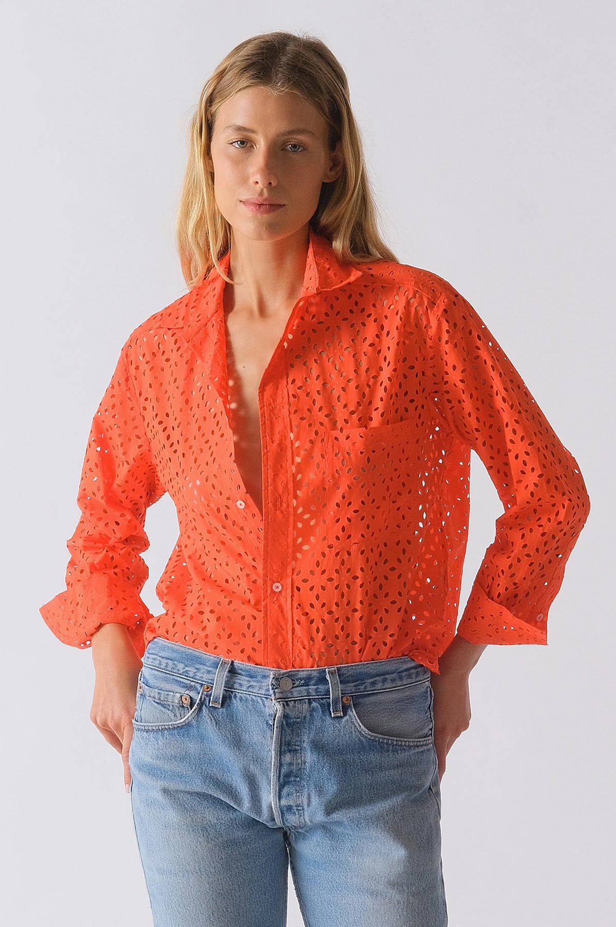 A woman wears The Daily Shirt by Cissa—a bright orange PAPER COTTON eyelet crop with rolled-up sleeves—paired with light blue jeans against a plain background, creating an effortless daily style statement.