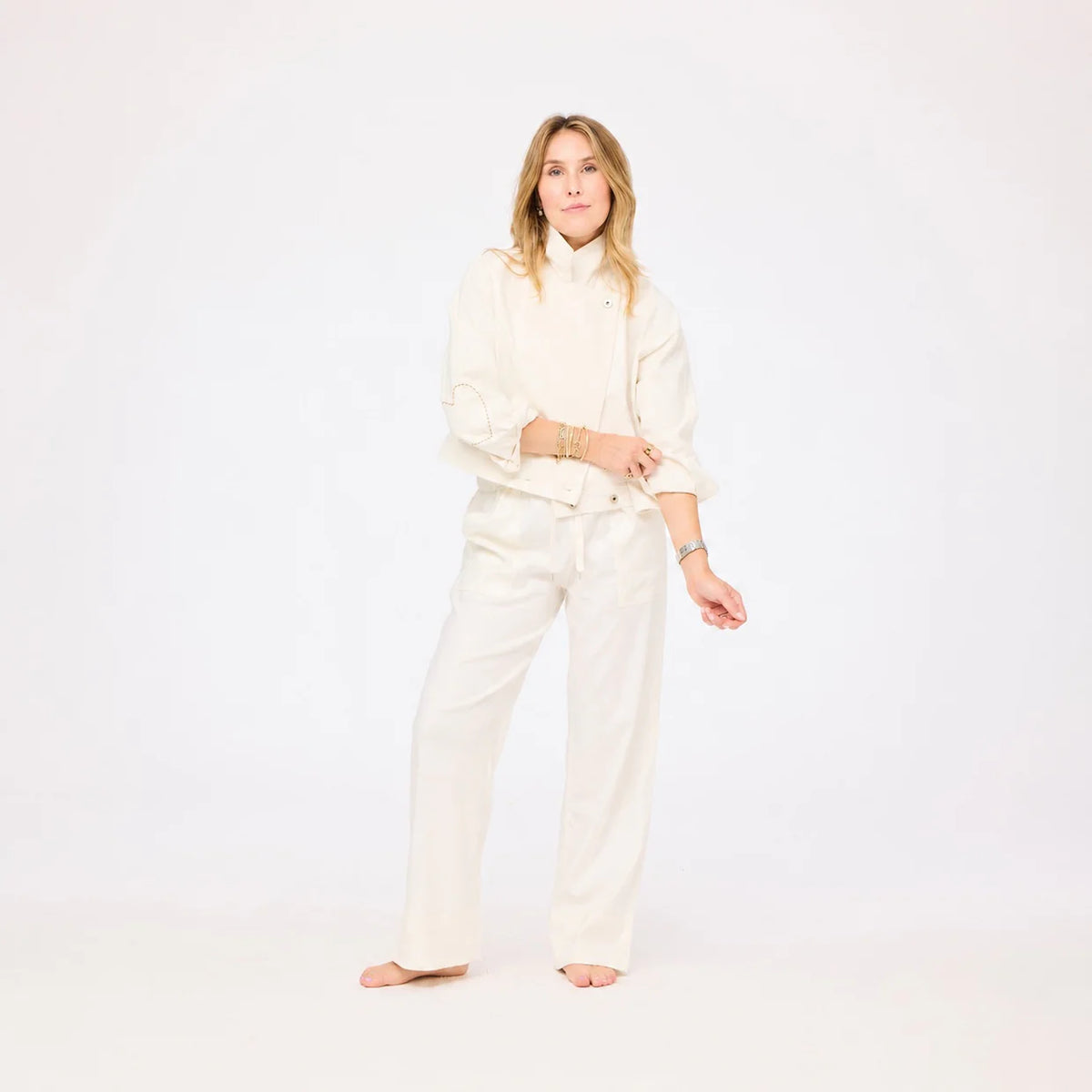A person with long, light brown hair stands barefoot on a white background, wearing the Kerri Rosenthal Denim Jacket in Cloud styled with wide-leg white denim pants and rolled sleeves.