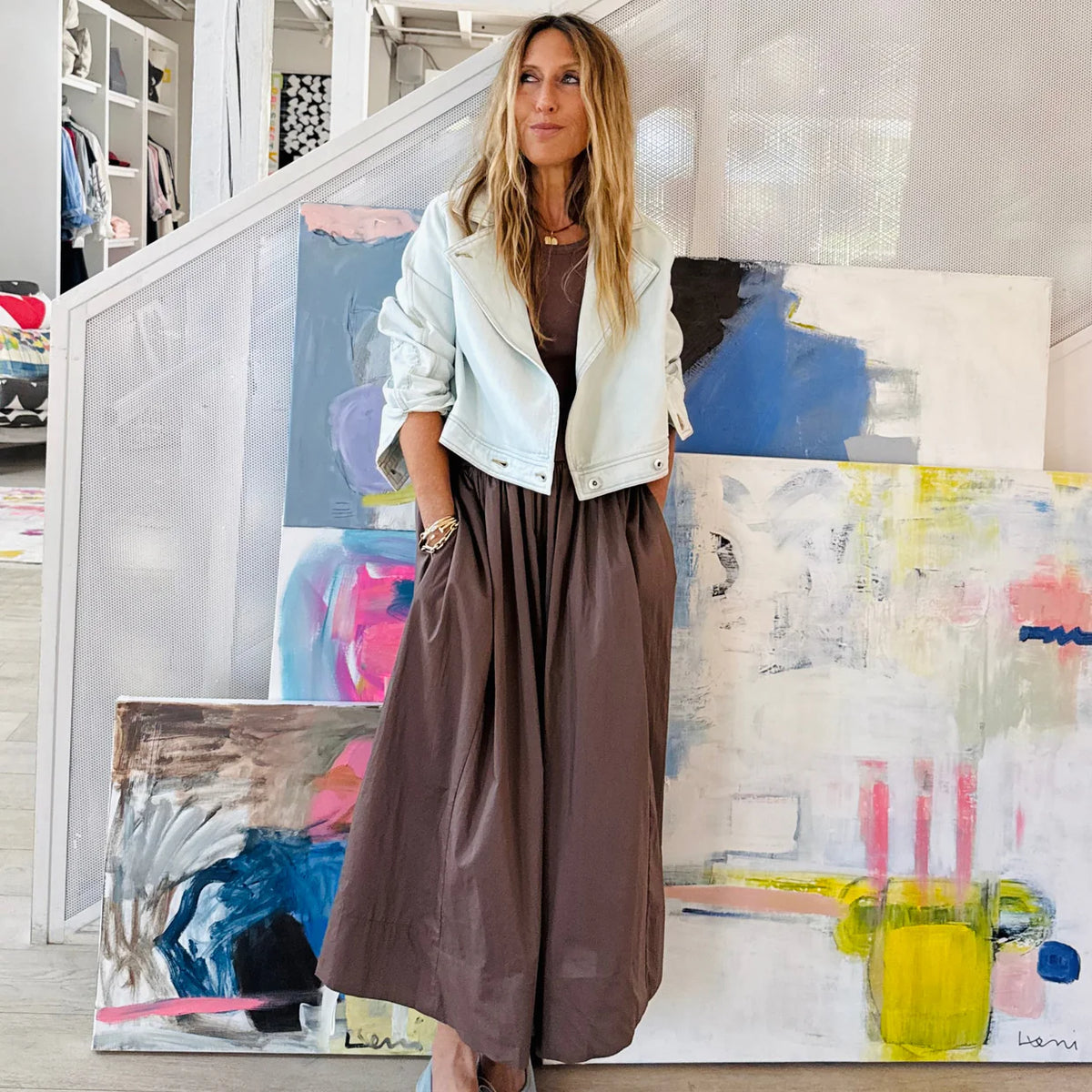 A woman wears the KR Tank Dress by Kerri Rosenthal with a light jacket, leaning against a wall indoors. Large colorful abstract paintings enhance the modern, artistic vibe of the setting.