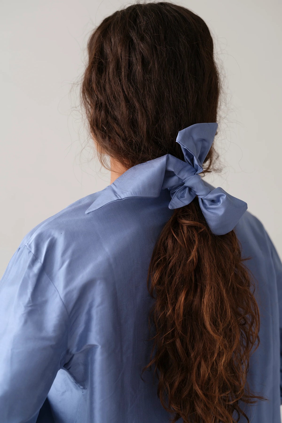 A person with long, wavy brown hair is seen from behind, wearing a light blue shirt and securing their ponytail with Donni's The Taffeta Bow Cornflower Blue accessory.