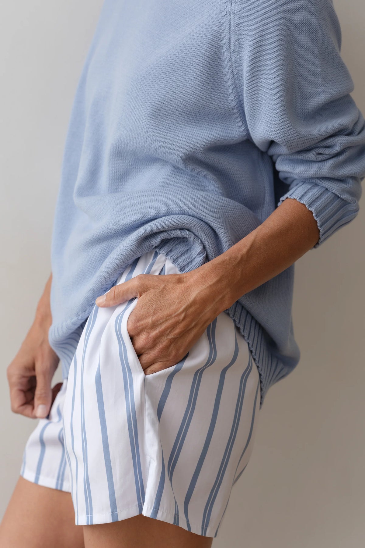 A person wearing The Pop Boxer shorts by Donni, featuring blue vertical stripes on white, and a light blue sweater, exudes a relaxed fit with one hand tucked in the pocket. Made from cotton poplin, they stand effortlessly against a plain light background.