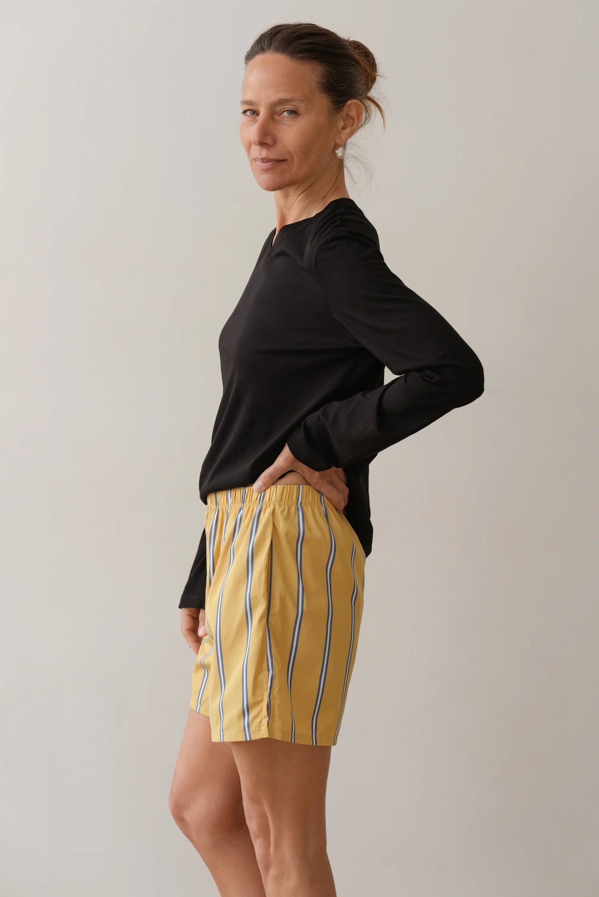 With their hair tied back, a person stands sideways and looks at the camera, wearing Donni's The Pop Boxer shorts in yellow with blue and white stripes, alongside a relaxed fit black long-sleeve shirt. They rest one hand on their hip against a plain, light backdrop.