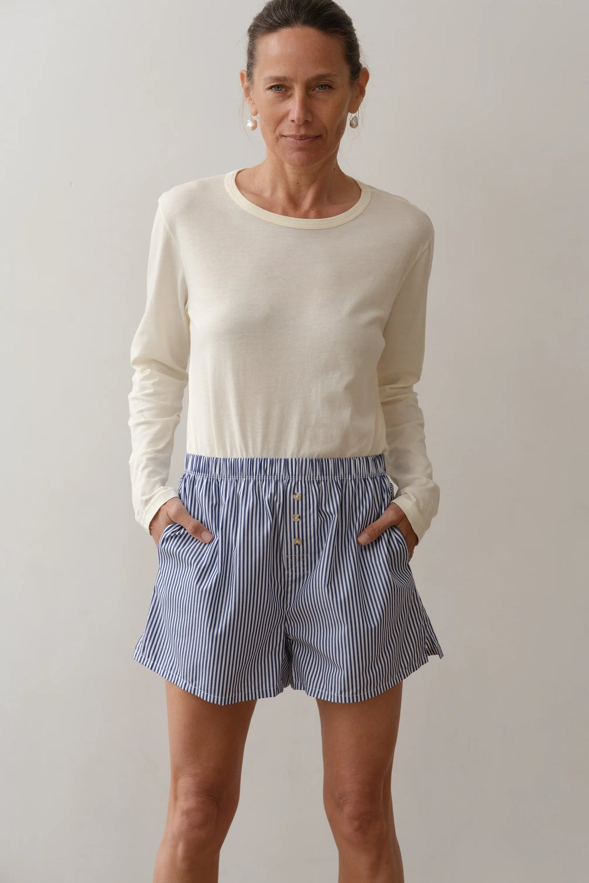 A woman stands against a plain background, wearing Donni's The Stripe Pop Boxer—blue-and-white striped cotton poplin boxers with buttons and pockets—and a long-sleeve cream shirt. She smiles slightly at the camera. Made in Los Angeles.