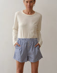 A woman stands against a plain background, wearing Donni's The Stripe Pop Boxer—blue-and-white striped cotton poplin boxers with buttons and pockets—and a long-sleeve cream shirt. She smiles slightly at the camera. Made in Los Angeles.