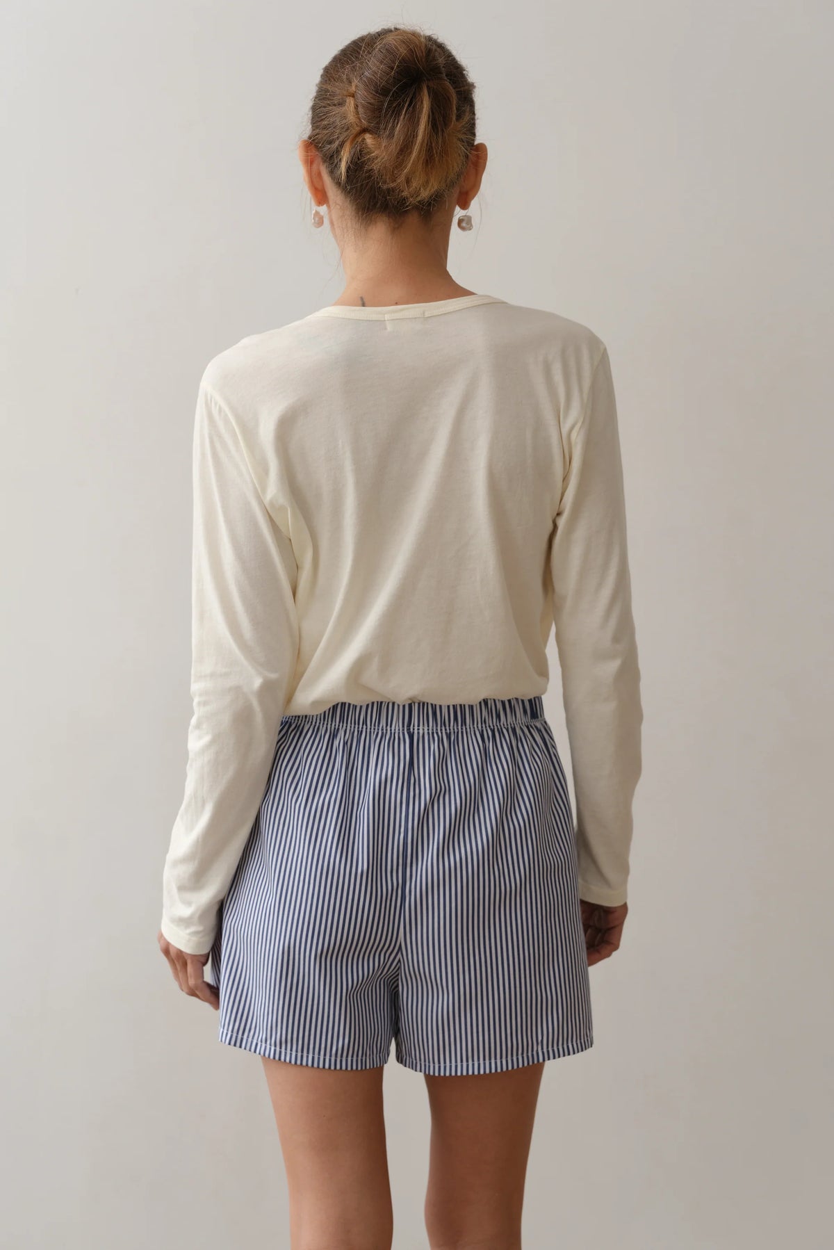 A person stands with their back to the camera, wearing Donni's The Stripe Pop Boxer—lightweight, breathable cotton poplin shorts with blue-and-white vertical stripes—paired with a long-sleeve cream shirt. Their hair is in a bun against a plain background.