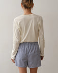 A person stands with their back to the camera, wearing Donni's The Stripe Pop Boxer—lightweight, breathable cotton poplin shorts with blue-and-white vertical stripes—paired with a long-sleeve cream shirt. Their hair is in a bun against a plain background.