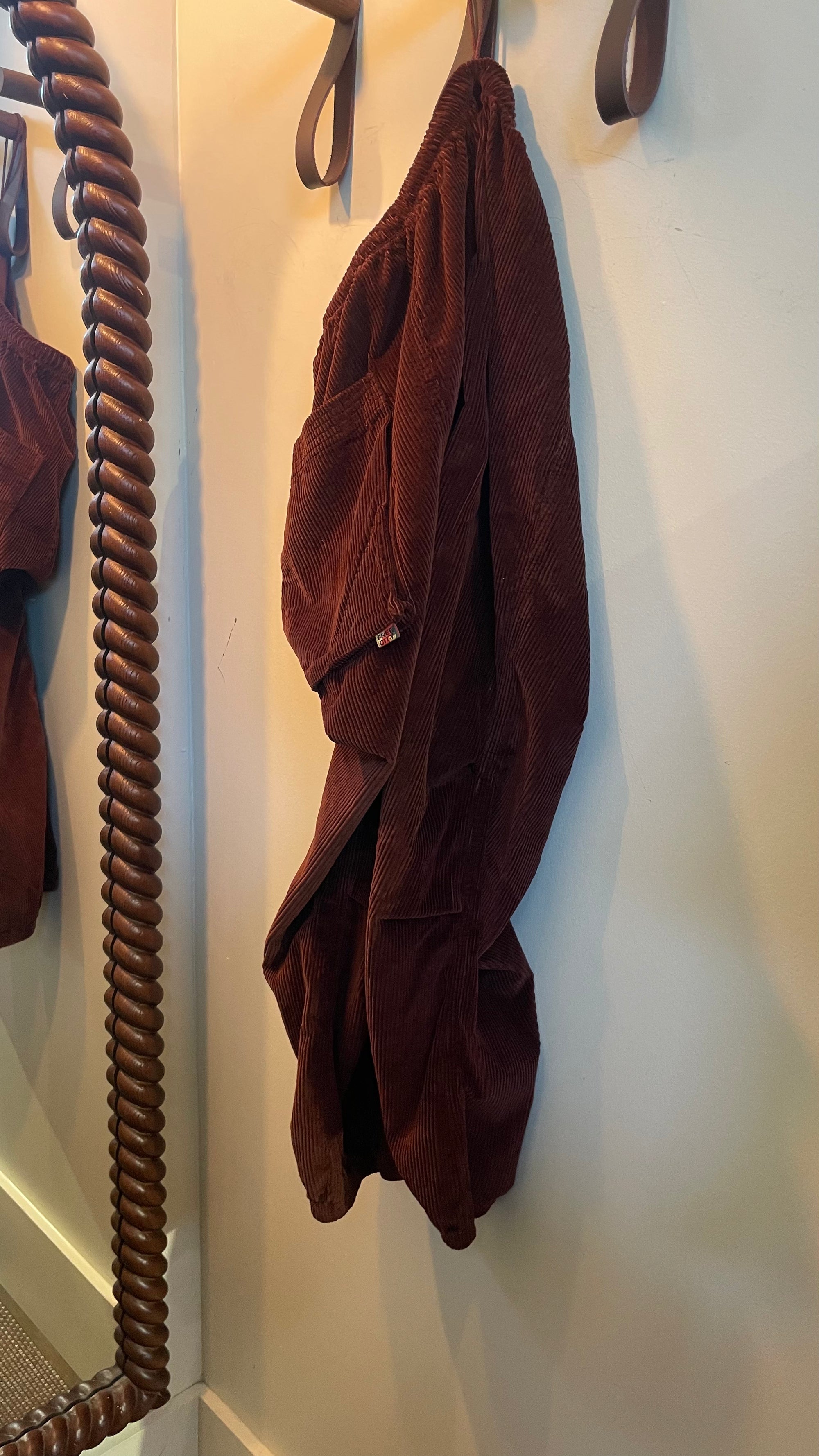 Free City Biggy corduroy pants in dark brown, hanging by a spiral-carved mirror frame.