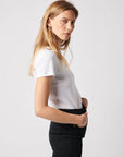 A woman with long blonde hair wears a white Majestic Filatures Deluxe Cotton S/S Crewneck tee and black pants, standing in profile against a plain light background, looking seriously at the camera.