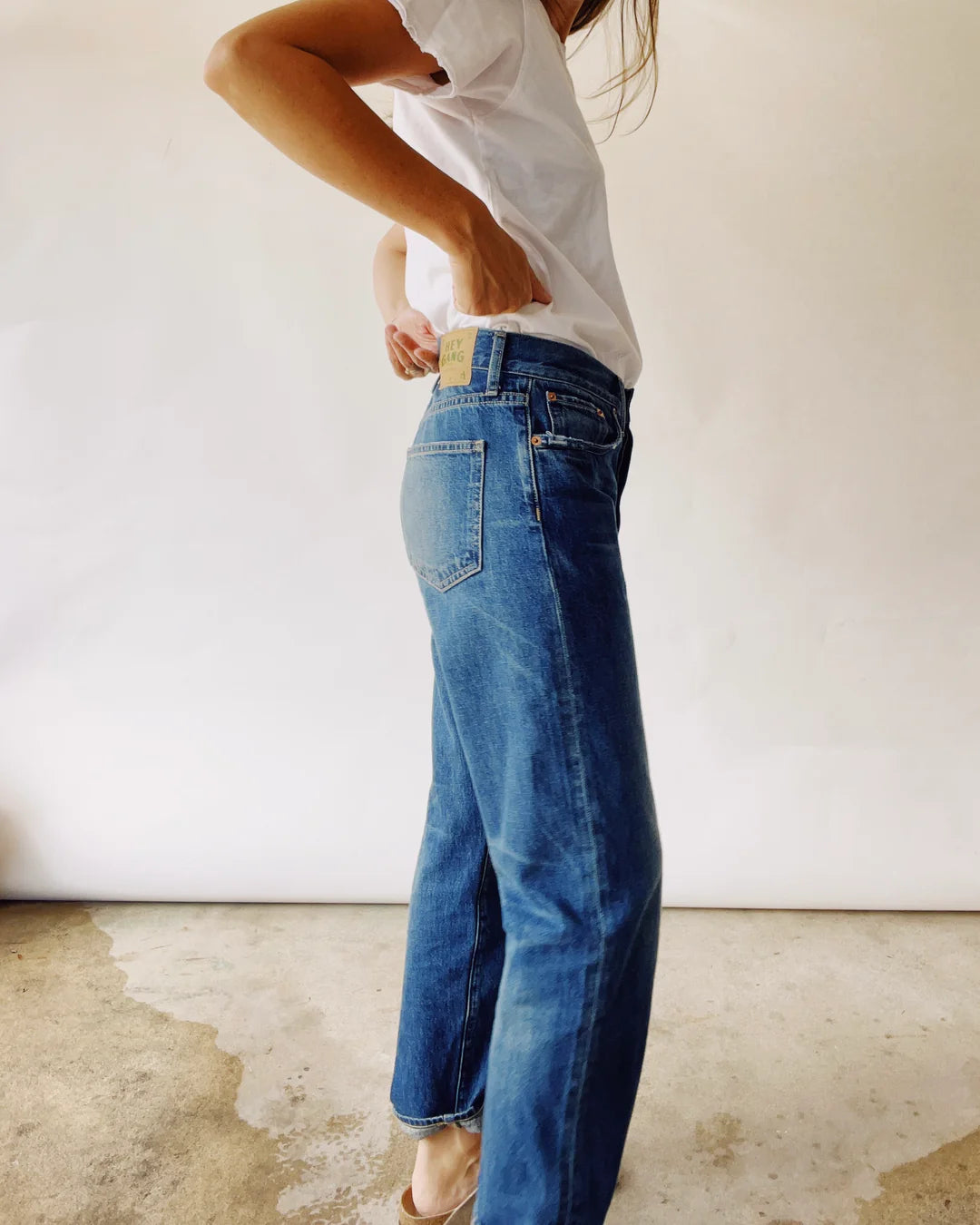 A person in a white t-shirt models Hey Gang's "The Loose Crop," relaxed mid-rise jeans made from 100% cotton denim. Standing on a textured floor, the side view highlights the jeans' fit and style against a plain backdrop.