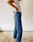 A person in a white t-shirt models Hey Gang's "The Loose Crop," relaxed mid-rise jeans made from 100% cotton denim. Standing on a textured floor, the side view highlights the jeans' fit and style against a plain backdrop.