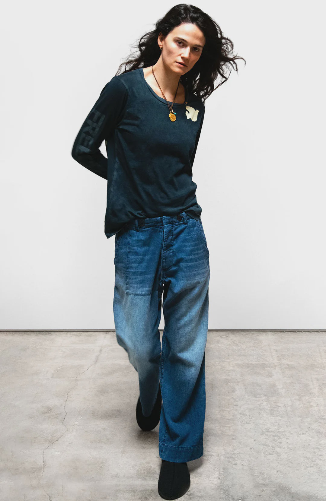 A person with long dark hair wears a black long-sleeve shirt, Free City Indigo French Sailor Pant, and black shoes, standing on a concrete floor against a plain white background.