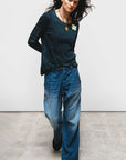 A person with long dark hair wears a black long-sleeve shirt, Free City Indigo French Sailor Pant, and black shoes, standing on a concrete floor against a plain white background.