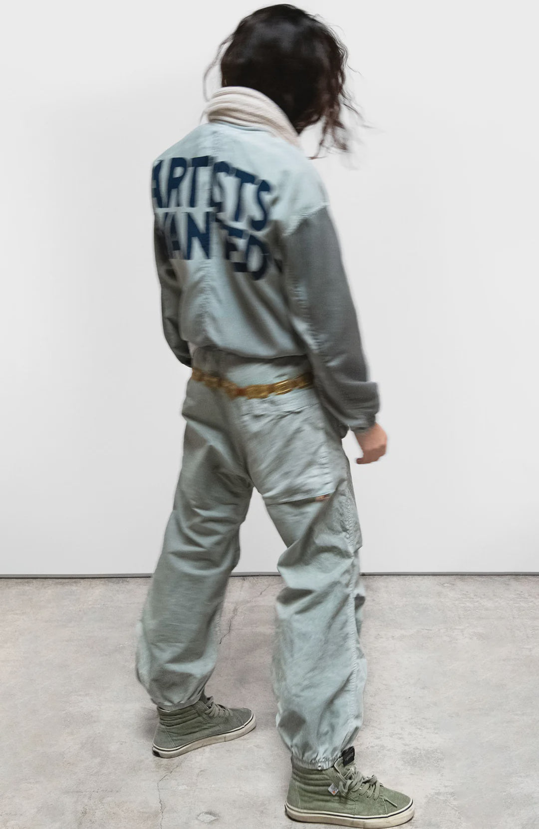 A person with wavy dark hair wears Free City's Flap/Snap AirJump Pant in light gray with a custom sunfade wash and "ARTISTS WANTED" on the back, green high-top sneakers, and a white scarf, standing on concrete facing away from the camera.
