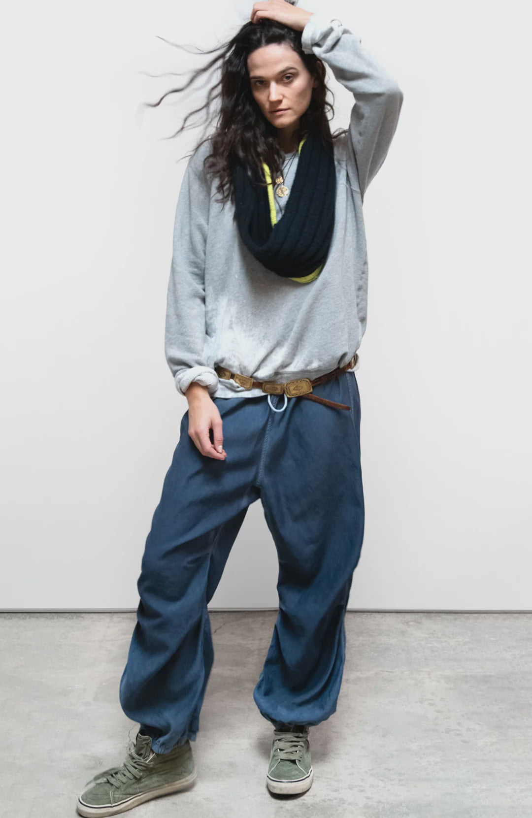 A person poses confidently against a plain background, wearing a loose gray sweatshirt, Free City (sparrow, LLC) Matte/Satin Biggy Pant in an oversized fit, olive green high-top sneakers, a belt, and has dark wavy hair framing their face.